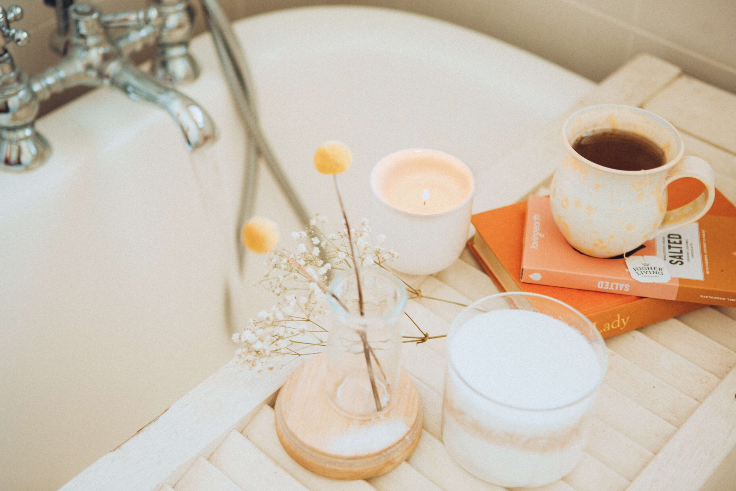 a close up shot of a bath being prepared for a night of self-care. on the table across the bathtub there is a ceramic mug of tea, a candle, an epsom salt container, and a little vase of picked wildflowers.