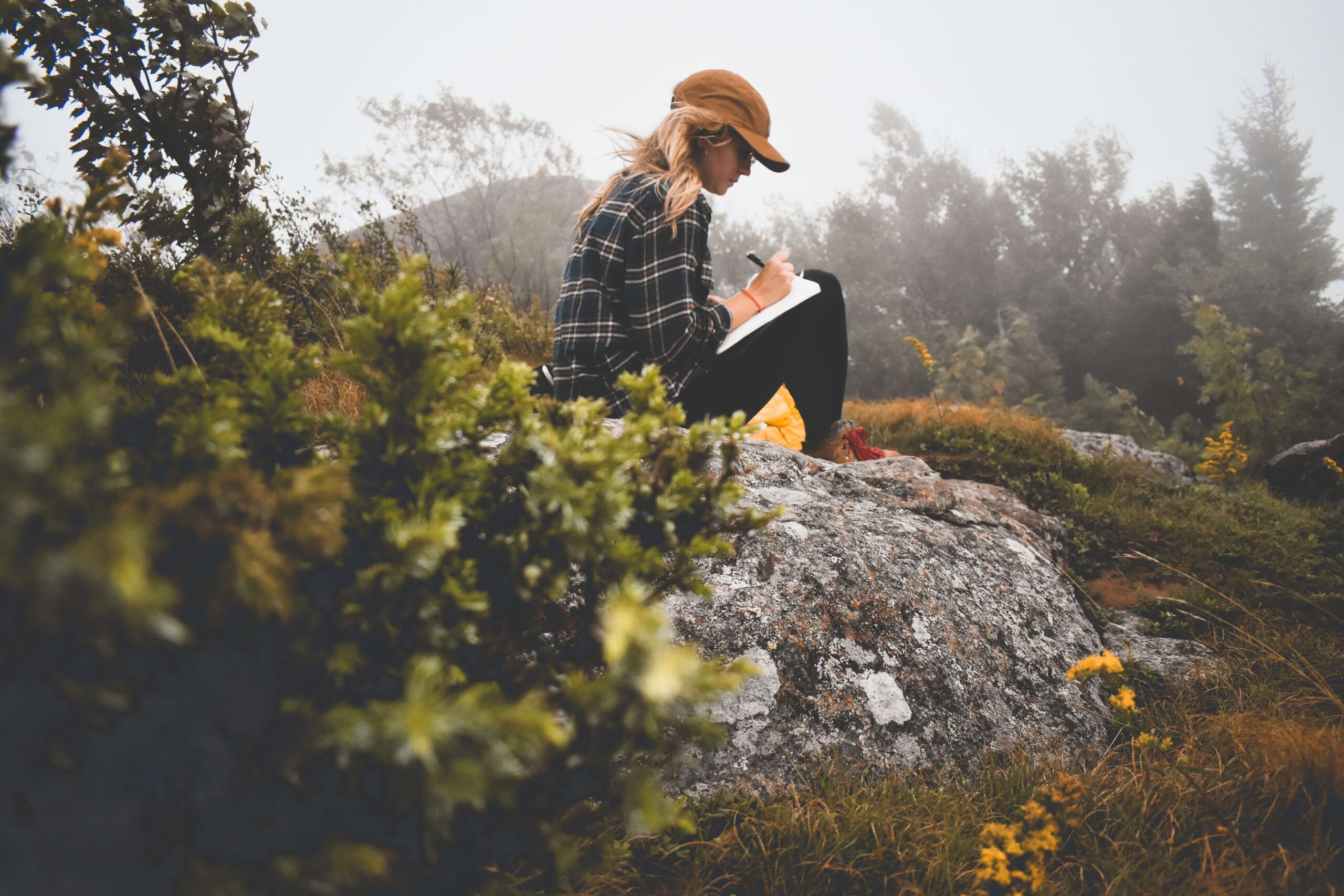 woman sitting on a rock on a hilltop journaling. she is wearing a mustard colored ball cap, plaid flannel shirt, black leggings, and brown hiking shoes with red laces. side profile view