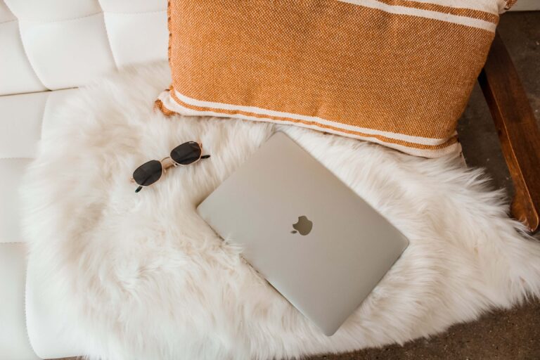 computer laying on white fur blanket with an orange pillow and sunglasses next to it while working from home